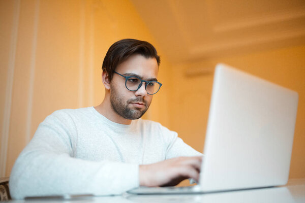 Serious young man working on laptop over yellow background. Dressed in white sweater and wearing blue glasses