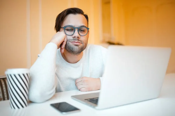 Stock image Tired businessman with laptop, smartphone and coffee on white table. Wearing blue glasses and sweater, over yellow background