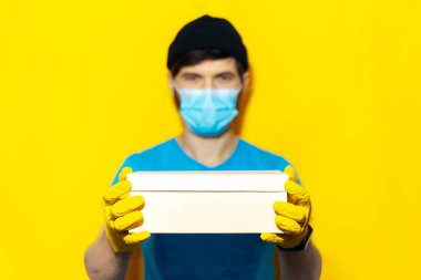 Coronavirus 'a karşı tıbbi maske ve eldiven takan teslimatçının elindeki karton kutuya yakın çekim. Sarı arkaplanda izole edilmiş.