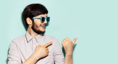 Güneş gözlüğü takan gülümseyen genç adamın stüdyo portresi, parmaklarıyla fotokopi alanı olan aqua menisinin boş arkaplanını gösteriyor..