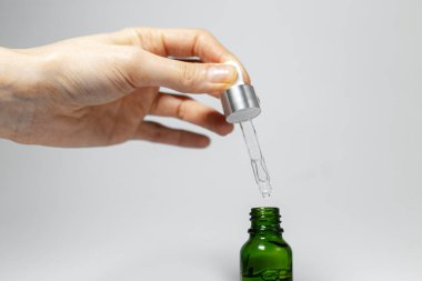 Yakın plan çekimde, temel cilt bakım yağı pipeti küçük cam bir şişe yeşile düşer. Grinin stüdyo arkaplanı.