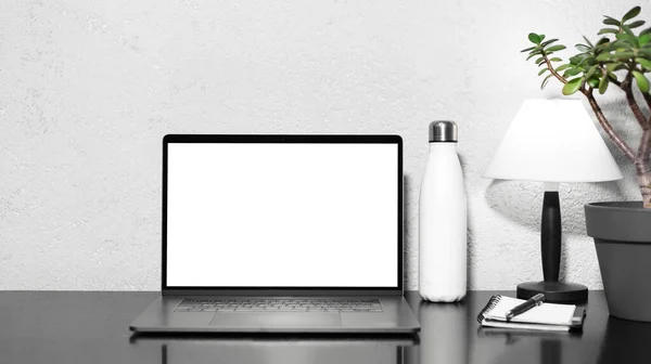 Serbest iş yeri olarak. Çelik termo su şişesi, lamba, çiçek ve kalemle birlikte defter yanında siyah masanın üstünde duran dizüstü bilgisayarı. Desenli duvar arkaplanı.