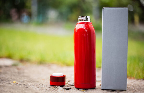Close-up of reusable steel red thermo bottle for water beside cap and cardboard box for packaging. Blurred outdoors background.