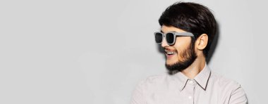 Güneş gözlüğü takan mutlu bir gencin stüdyo portresi. Fotokopi alanı olan gri rengin boş arka planına bakıyor..