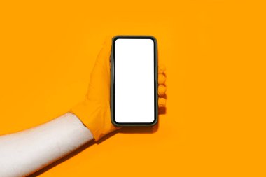Turuncu eldiven giyen ve turuncu arka planda akıllı telefon tutan erkek eli yakın plan..