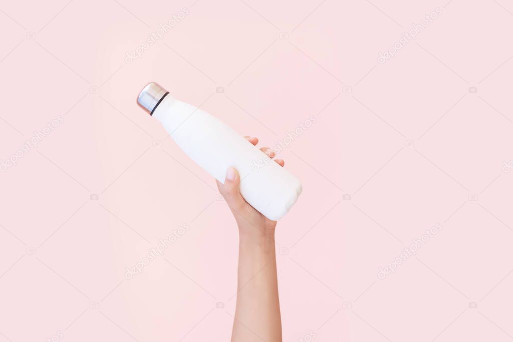 Close-up of female hand holding reusable, steel eco thermo water bottle of white, isolated on background of pastel pink color. Be plastic free. Zero waste. Environment concept.