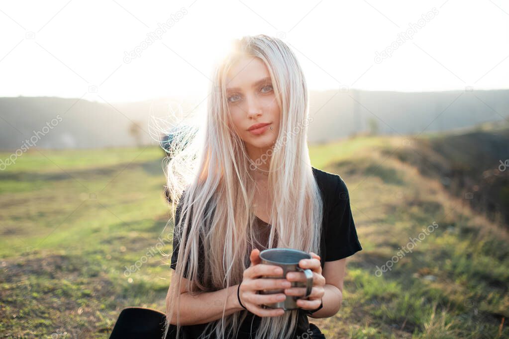 Portrait of young beautiful blonde girl, holding a warm steel cup with tea on the background of sunset.