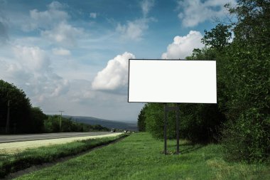 Asfaltlanan yol ve yeşil ormanın yakınındaki reklam afişi için boş ilan panosu, mavi gökyüzünün arka planı..