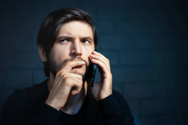 Close-up portrait of young thoughtful man, talking on smartphone. Background of black brick wall.