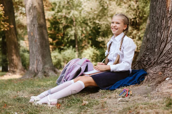 A schoolgirl does lessons in a park under a tree .schoolgirl sits under a tree in the park