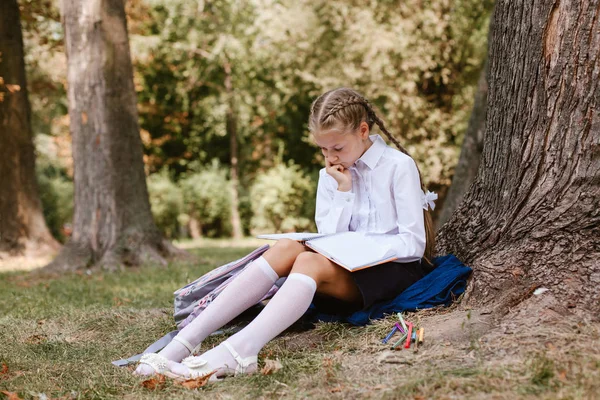 A schoolgirl does lessons in a park under a tree .schoolgirl sits under a tree in the park