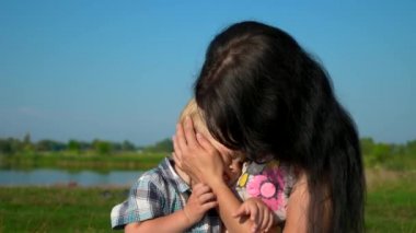 Mutlu genç sarılma ve öpüşme şirin çocuk ellerinde tutan anne. Yaz nehir Gölü sabah güneşli gün mavi gökyüzü. Ağır çekim 30p 0,5 60 p