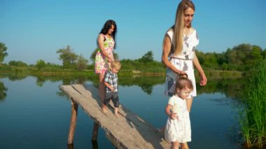 İki genç güzel anne yürüyüş ve su onların çocukları ile oynamak. Yaz nehir sabah güneşli gün mavi gökyüzü. Ağır çekim 30p 0,5 60 p