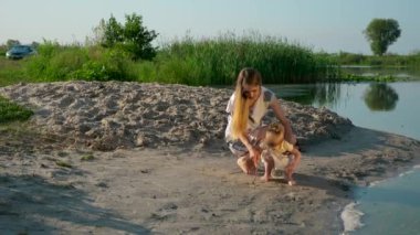  Genç güzel anne oyun çocuk ile. Nehir Lake yakınındaki kum üzerinde çizin. Yaz sabahı güneşli gün mavi gökyüzü. Yavaş hareket 30 p 0,5 60 p