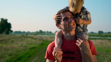 Genç yakışıklı tatlı çocuk omuzlarında taşıyan Baba adam. Yaz sabahı güneşli bir gün mavi Clea gökyüzü kırsal yol. Yavaş hareket 30 p 0,5 60 p