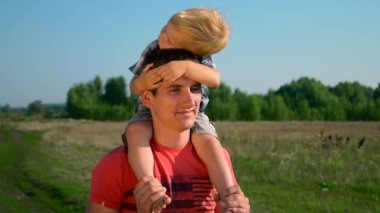 Genç yakışıklı tatlı çocuk omuzlarında taşıyan Baba adam. Yaz sabahı güneşli bir gün mavi Clea gökyüzü kırsal yol. Yavaş hareket 30 p 0,5 60 p