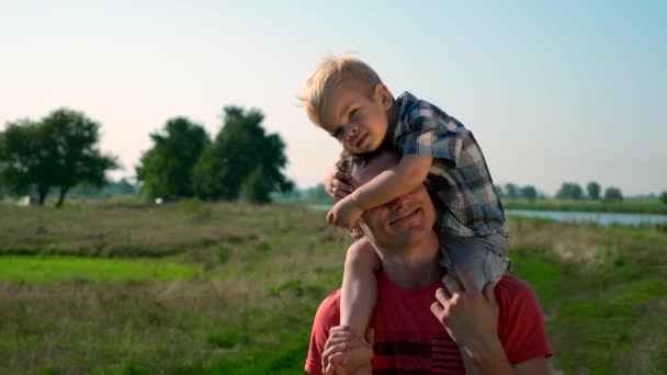 ハンサムな若者の肩の上のかわいい子を運ぶ父 夏の朝晴れた青空クレア田舎道 スローモーション — ストック動画