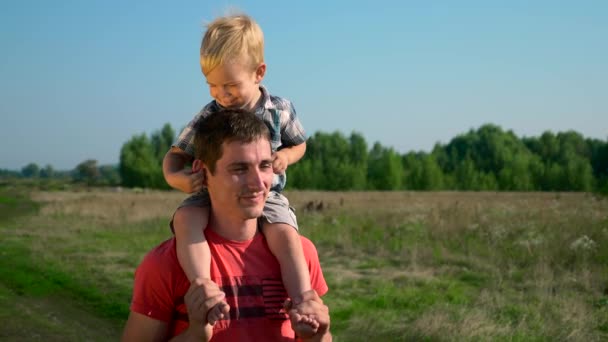 ハンサムな若者の肩の上のかわいい子を運ぶ父 夏の朝晴れた青空クレア田舎道 スローモーション — ストック動画
