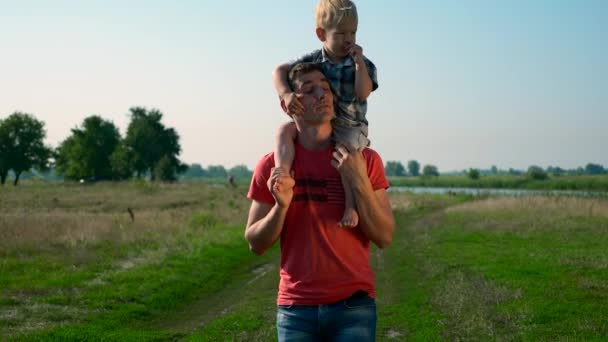 年轻英俊的男子父亲背着可爱的孩子的肩膀上 夏日清晨阳光明媚的一天 蓝天晴朗的乡间小路 慢动作 30P 60P — 图库视频影像
