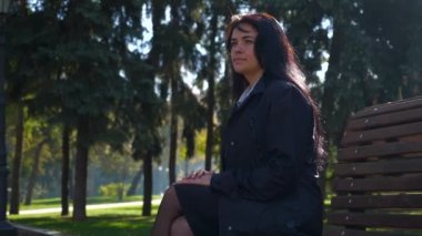Bankta oturan çekici güzel kız yerleşmek Şehir Parkı ve düşünme. Sonbahar güneşli gün. Yavaş hareket 4k 60 fps