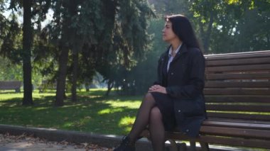 Bankta oturan çekici güzel kız yerleşmek Şehir Parkı ve düşünme. Sonbahar güneşli gün. Yavaş hareket 4k 60 fps