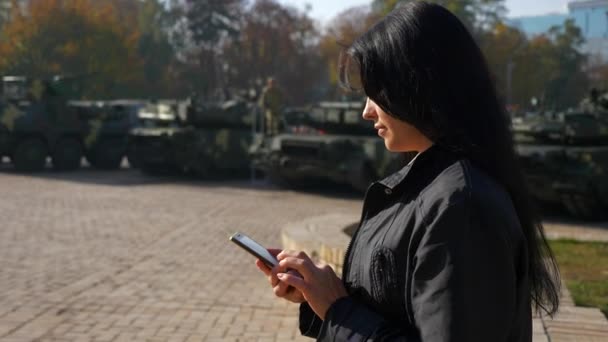 美丽的褐发女郎使用智能手机 背景下城市广场上的军车 慢动作 2019年乌克兰基辅 — 图库视频影像