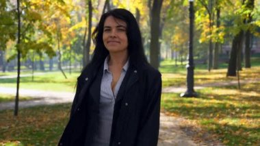 Kamera izler gülümser ve güzel ciddi kız görünüyor geri yürüme şehir parkı ve düşünme. Şehir Parkı sabah. Sonbahar güneşli gün. 4k yavaş hareket 60 fps