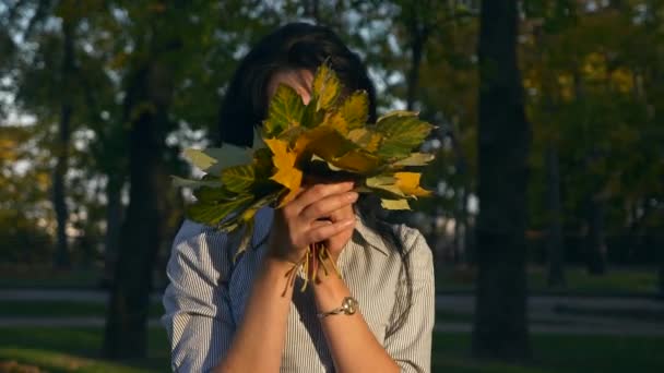 美丽的女孩把叶子握在手里 转身躲在树叶后面 快乐的积极情绪 晚上黄金时间在公园里 慢动作 Fps — 图库视频影像