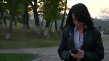 İşten sonra dönen. Akşam şehir Park'ta yürüyordunuz akıllı telefon kullanan güzel kız. Akşam (sabah) şehir kentsel arka plan. Yavaş hareket 4k 60fps