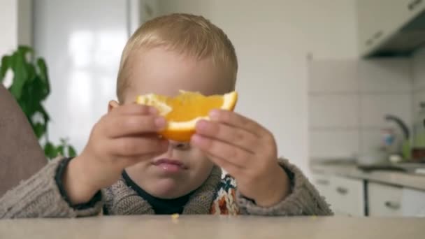 のかわいい小さな男の子は台所でおいしいオレンジを食べる 背景の親 忙しい母の朝食夕食 — ストック動画