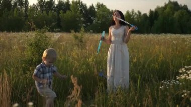 Genç Anne Sabun Baloncukları Üflüyor ve Küçük Neşeli Oğul Yüksek Çimen Tarlasında Yakalıyor. Akşam Günbatımı Kırsal Sahne. 2x Slow motion - Yarı hız 60 FPS