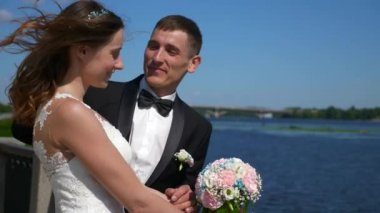 Gelin ve Damat, Nehir kenarındaki Quay 'da duruyor. Rüzgarlı Düğün Günü. El sallayan saç. Şehir manzarası Şehir Sahne Arkaplanı. 2x Yavaş çekim - 0.5 Hız 60 FPS