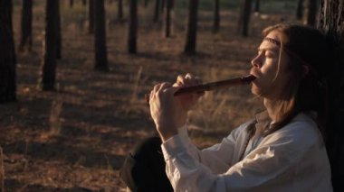 Uzun saçlı adam çam ağaçlarının altında flüt çalar. Çelenkli Arka plan Kadını ve Ormanda Sepet Yürüyüşü. Yavaş çekim 60 FPS