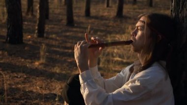 Uzun saçlı adam çam ağaçlarının altında flüt çalar. Çelenkli Arka plan Kadını ve Ormanda Sepet Yürüyüşü. Yavaş çekim 60 FPS