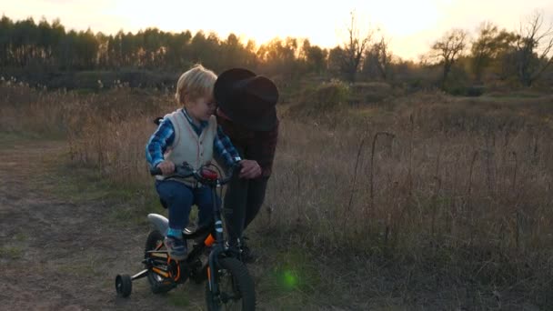 在教他骑婴儿车的同时 爱母亲会亲吻孩子 夕阳西下的乡村风景 慢动作 — 图库视频影像