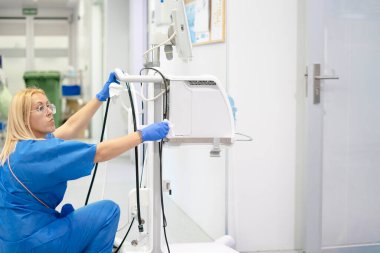 Female doctor wearing blue scrubs and gloves cleaning medical equipment in hospital room