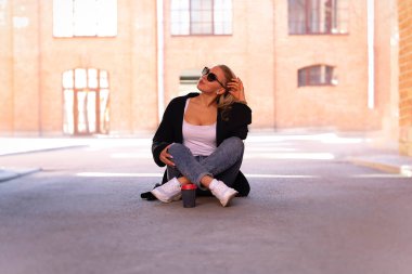 Uzun saçlı, kot pantolonlu, beyaz kolsuz tişörtlü, siyah kazaklı ve beyaz ayakkabılı bir sarışın. Kaldırım kenarında dinleniyor. Ayakta bir fincan kahve var.