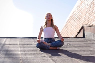 Sarı saçlı bir kız çatıda meditasyon yapmak için inzivaya çekilmiş. Beyaz tişört ve kot pantolon giymişti.