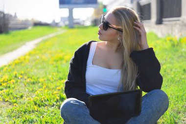 Çimlerin üzerinde oturup dizüstü bilgisayarda çalışırken çok düşünceli bir sarışın. Siyah kazak, kurşun tişört ve kot pantolon giymişti. Gözlere güneş gözlüğü.