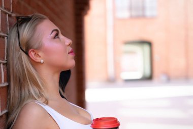 Sarışın, sırtı tuğla duvara dönük duruyor. Bir bardak kahvenin ellerinde.