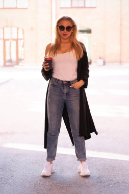 Güzel bir sarışın kemerde duruyor. Elinde kahve tutuyordu. Güneş gözlüklerinin önünde. Arka planda kavurucu güneş