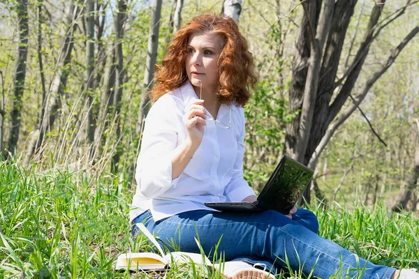 Kızıl saçlı kız elinde gözlük tuttuğunu sanıyordu. Çevrimiçi iş. Çimenler, ağaçlar.