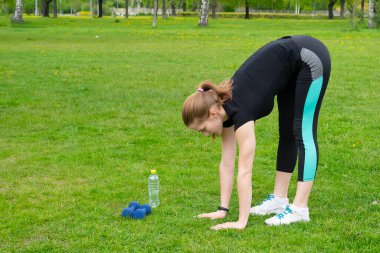 Kız, parkta fitness ile meşgul. Öne eğiliyor ve elleriyle yere uzanıyor. Yakınlarda bir şişe su ve dambıl var..