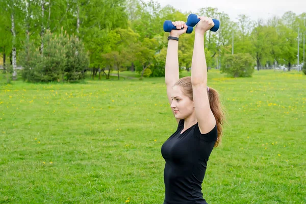 beautiful girl is engaged in fitness in the park near the house. Understands dumbbell up. On hand fitness bracelet