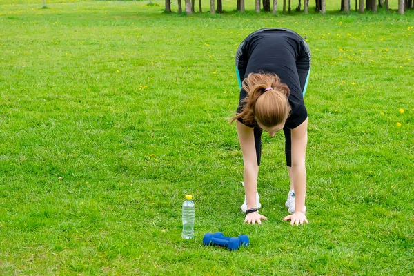 Güzel bir kadın doğada spor yapmakla meşgul. Eğiliyor. Çimenlerin üzerinde halterler ve bir şişe kemer var.