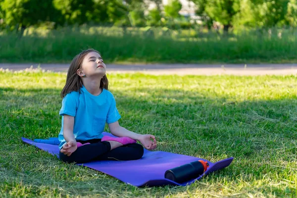 Küçük kız parkta halının üzerinde oturarak yoga yapıyor. Lotus konumu