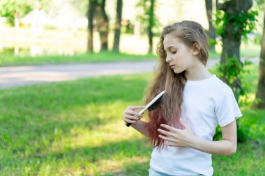 Küçük kız saçını tarıyor. Parktaki çimlerin üzerinde duruyor. Ağaçlara karşı..