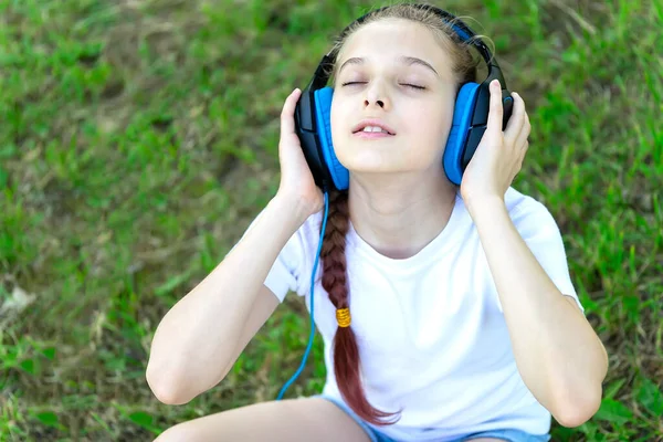 Parkta çimlerin üzerinde kulaklıkla müzik dinleyen küçük bir kız. Ön görünüm