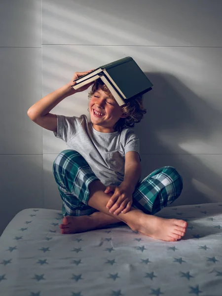 Pijamalı Sevimli Bir Çocuk Kafasına Kitap Takmış Rahat Bir Yatakta — Stok fotoğraf