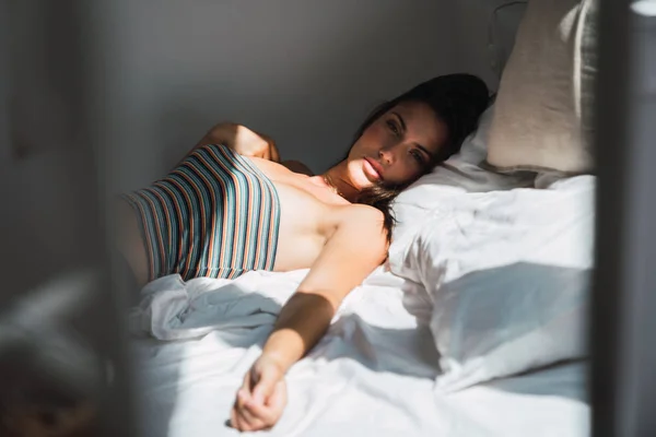 年轻的沉思的女人在条纹作物顶躺在床上 在阴影的卧室 看着相机 — 图库照片